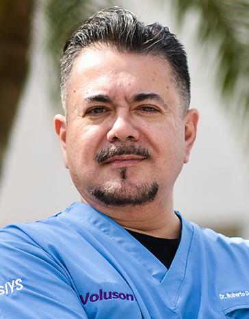 Es la foto de perfil de un ginecólogo y obstetra en Villahermosa de Hospital Angeles Villahermosa. Usa un uniforme médico azul. Tiene el cabello y barba negro. Está sonriendo ligeremante.