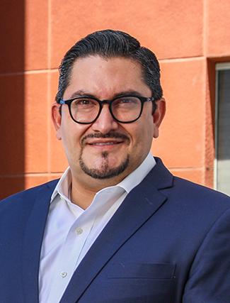 Es la foto de perfil de un urólogo en León de Hospital Angeles León. Tiene el cabello y barba negra. Usa lentes y un saco azul marino con una camisa azul claro.