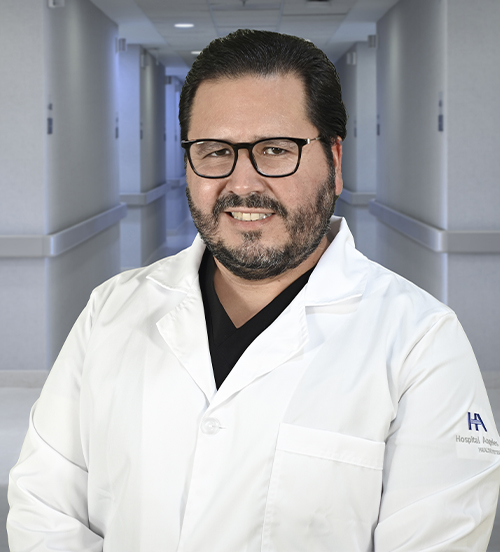 Es la foto de perfil de un radiólogo en Ciudad Juárez de Hospital Angeles Ciudad Juárez. El doctor tiene barba y bigote, cabello oscuro y corto, y lleva gafas. Está sonriendo y viste un uniforme médico de color azul marino con un pequeño logotipo del hospital en el pecho. El fondo muestra una pintura en tonos cálidos con figuras abstractas, creando un ambiente acogedor.