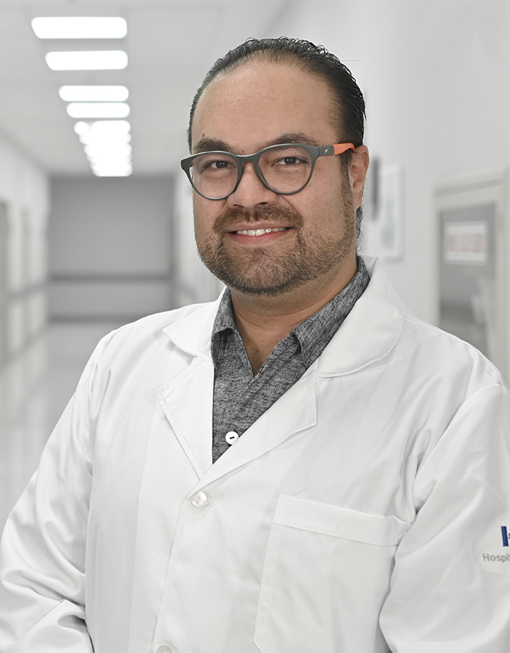 Es la foto de perfil de un ortopedista y traumatólogo en Ciudad de México de Hospital Angeles Universidad. Usa una bata blanca con una camisa gris. Tiene el cabello y barba café oscuro. Usa lentes y está sonriendo.