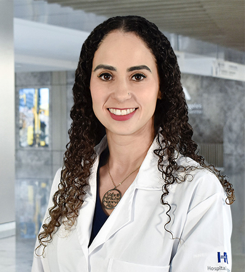 Es la foto de perfil de una nutrióloga en Querétaro de Hospital Angeles Centro-sur. Tiene el cabello color café y chino. Usa una bata blanca en conjunto de un uniforme médico azul oscuro. Está sonriendo y usa un collar con un dije grande.