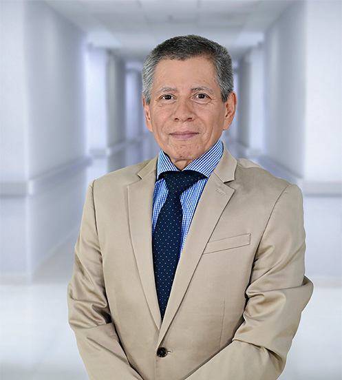 Es la foto de perfil de un anestesiólogo en Ciudad Juárez de Hospital Angeles Ciudad Juárez. El doctor tiene cabello canoso y lacio, peinado hacia un lado. Está sonriendo ligeramente y lleva un traje oscuro con corbata azul marino con pequeños detalles, junto con una camisa blanca. El fondo muestra un área luminosa del hospital, con paredes claras y una pintura en tonos cálidos.