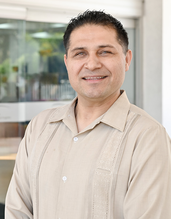Es la foto de perfil de un hematólogo en Tijuana de Hospital Angeles Tijuana. Usa una bata blanca con una camisa gris. Tiene el cabello y barba razurada negra. Está sonriendo ligeramente.