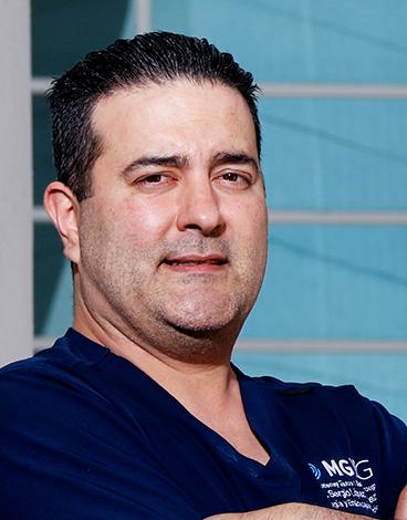 Es la foto de perfil de un especialista en endoscopía y cirujano general en San Pedro Garza de Hospital Angeles Valle Oriente. Usa un uniforme médico azul. Tiene el cabello negro. Tiene los brazos cruzados y está sonriendo ligeramente.