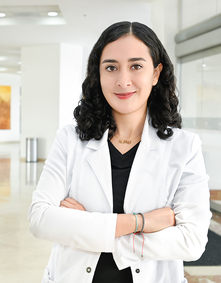Es la foto de perfil de un psiquiatra en Ciudad Juárez de Hospital Angeles Ciudad Juárez. La doctora tiene el cabello oscuro, rizado y suelto. Está sonriendo y viste una bata médica blanca sobre una camiseta negra. Lleva un collar dorado con un nombre grabado. El fondo muestra un área luminosa del hospital, con paredes claras y un diseño moderno.