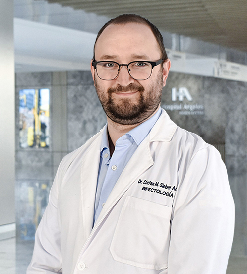 Es la foto de perfil de un infectólogo en Querétaro de Hospital Angeles Centro-sur. Usa una bata blanca con una camisa azul claro. Tiene el cabello y barba color castaño. Usa lentes.