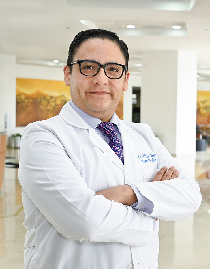 Es la foto de perfil de un cirujano oncológico en Ciudad Juárez de Hospital Angeles Ciudad Juárez. El doctor tiene el cabello oscuro, corto y peinado hacia atrás. Lleva gafas y está sonriendo ligeramente. Viste una bata médica blanca con su nombre y la especialidad 'Cirugía Oncológica