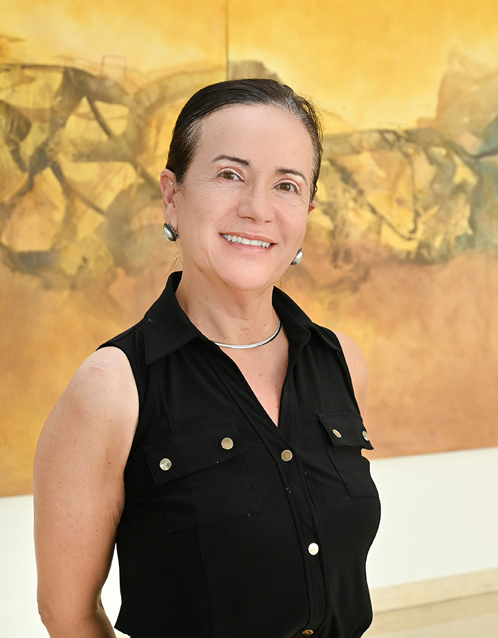 Es la foto de perfil de un ginecólogo y obstetra en Ciudad Juárez de Hospital Angeles Ciudad Juárez. La doctora tiene el cabello oscuro, peinado hacia atrás, y lleva aretes grandes de color plateado. Está sonriendo y viste una blusa sin mangas de color negro con botones. El fondo muestra una pintura en tonos cálidos con figuras abstractas.