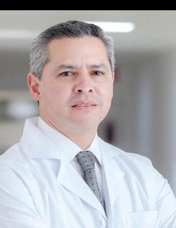 Es la foto de perfil de un cirujano neurológico en San Luis Potosí de Hospital Angeles San Luis Potosí. Usa una bata y camisa blanca con una corbata gris. Tiene el cabello gris. Está cruzado de brazos. Está serio.