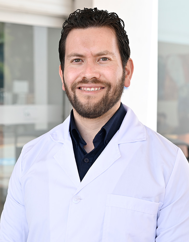 Es la foto de perfil de un urólogo en Tijuana de Hospital Angeles Tijuana. Usa una bata y camisa blanca y corbata lila. Tiene el cabello y barba castaño. Está sonriendo.