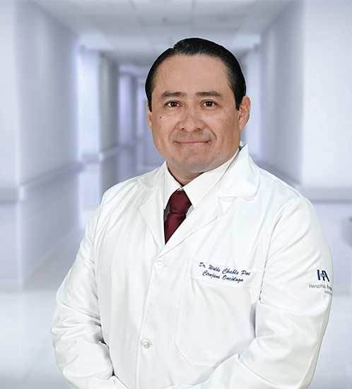 Es la foto de perfil de un cirujano oncológico en Ciudad Juárez de Hospital Angeles Ciudad Juárez. El doctor tiene el cabello oscuro y corto, peinado hacia atrás. Está sonriendo y lleva una bata médica blanca con su nombre y la especialidad 'Oncología Ginecológica' bordados en el pecho, junto con una corbata roja. En la manga de la bata se distingue el logotipo de 'Grupo Ángeles Servicios de Salud'. El fondo muestra un área luminosa del hospital, con paredes claras y una pintura en tonos cálidos.