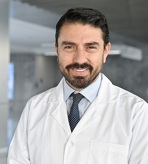 Es la foto de perfil de un neurólogo en Querétaro de Hospital Angeles Centro Sur. Usa una bata blanca, con una camisa blanca y una corbata morada con lunares blancos. Tiene barba y está sonriendo.